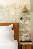 Una cama con sábanas blancas y un cabecero marrón, una mesita de noche redonda de madera con un objeto blanco y una pared con textura que tiene una lámpara colgante.