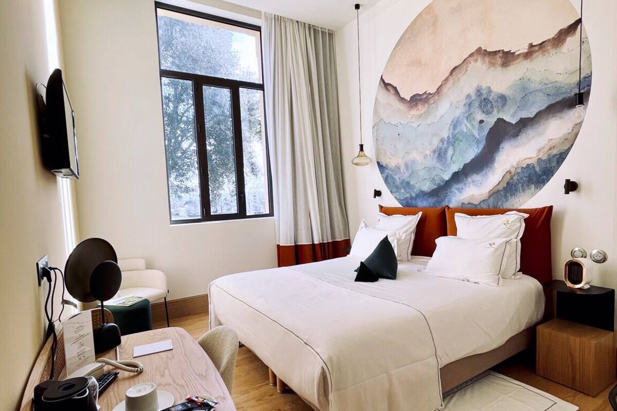A modern hotel room with a large bed, a desk with a chair, a large window, and a wall-mounted artwork.
