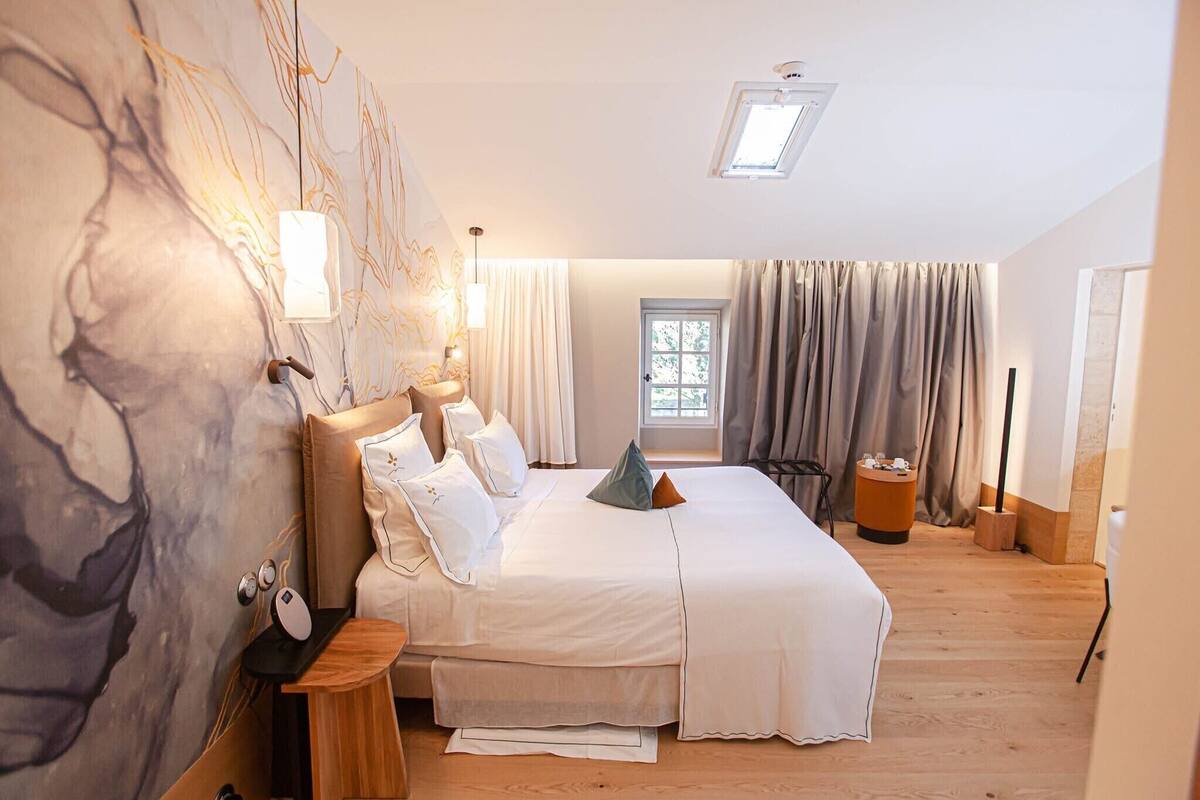 A bedroom with a large bed, a desk with a computer, a chair, a window with curtains, and a wall with a large abstract painting.