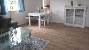 Ein modernes Wohnzimmer mit Glastisch, Esstisch mit Stühlen, einem weißen Schrank mit Glastüren, einer Wanduhr und einem Blumentopf.