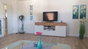 Ein modernes Wohnzimmer mit Glastisch, Flachbildfernseher auf einer Holzkommode, einem Topfpflanzen und einem Kunstwerk an der Wand.