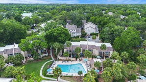 Vista aérea de una zona residencial con piscina, rodeada de palmeras y una casa grande de techo inclinado.