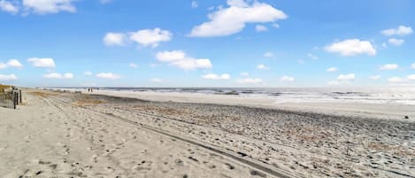 A beach with a sandy path leading to the water, a fence on the left, and a clear sky.