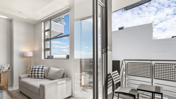 A modern living room with a sofa, a coffee table, and a chair. There is a large window offering a view of the sky and buildings. - Modern And Trendy 1 Bed Across From Rosebank Mall (Johannesburg)