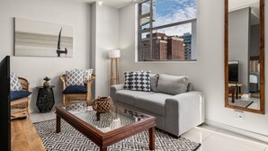 A modern living room with a grey sofa, a coffee table, wicker chairs, a large mirror, and a painting of a sailboat. - Modern And Trendy 1 Bed Across From Rosebank Mall (Johannesburg)