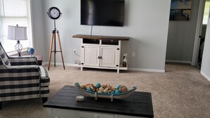 A living room with a flat-screen TV mounted on the wall, a tripod floor lamp, a sofa, a coffee table with decorative items, and a cabinet with drawers.
