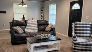 A living room with a black leather couch, a white coffee table, and a plaid armchair. A 'WELCOME' sign is on the wall, and there is a window with blinds.
