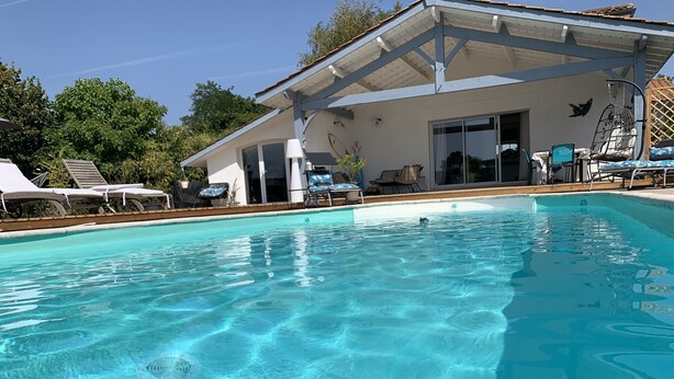 A clear swimming pool with lounge chairs and umbrellas, a covered patio area, and a building with large windows and doors. - Villa 5 Piéces Avec Piscine (Le Porge)
