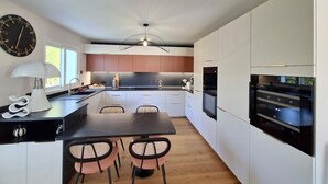 A modern kitchen with a central island, white cabinetry, a black countertop, and a dining area with two chairs. - Villa 5 Piéces Avec Piscine (Le Porge)