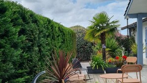 A patio area with a seating arrangement, a tall green hedge, and a variety of plants. - Villa 5 Piéces Avec Piscine (Le Porge)