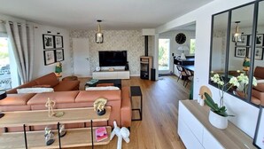 A modern living room with a sofa, TV, and a dining area with chairs and a table. - Villa 5 Piéces Avec Piscine (Le Porge)