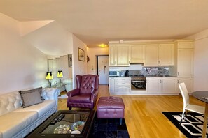 Una sala de estar moderna con un sofá blanco, un sillón de cuero y una mesa de centro de vidrio. Se ve una cocina con armarios blancos y una zona de comedor con una silla blanca al fondo.