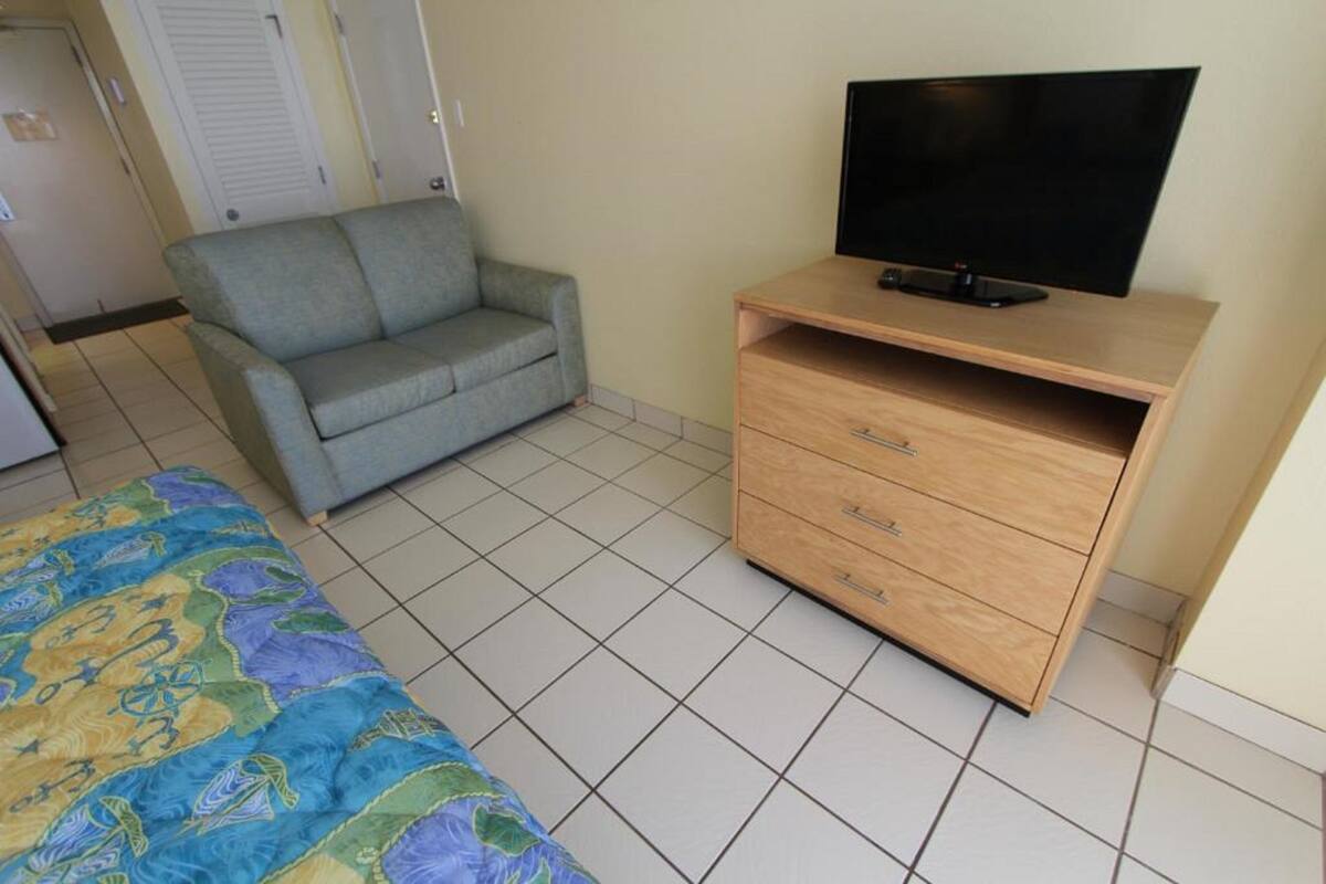 A room with a bed, a grey sofa, a wooden TV stand, and a tiled floor.
