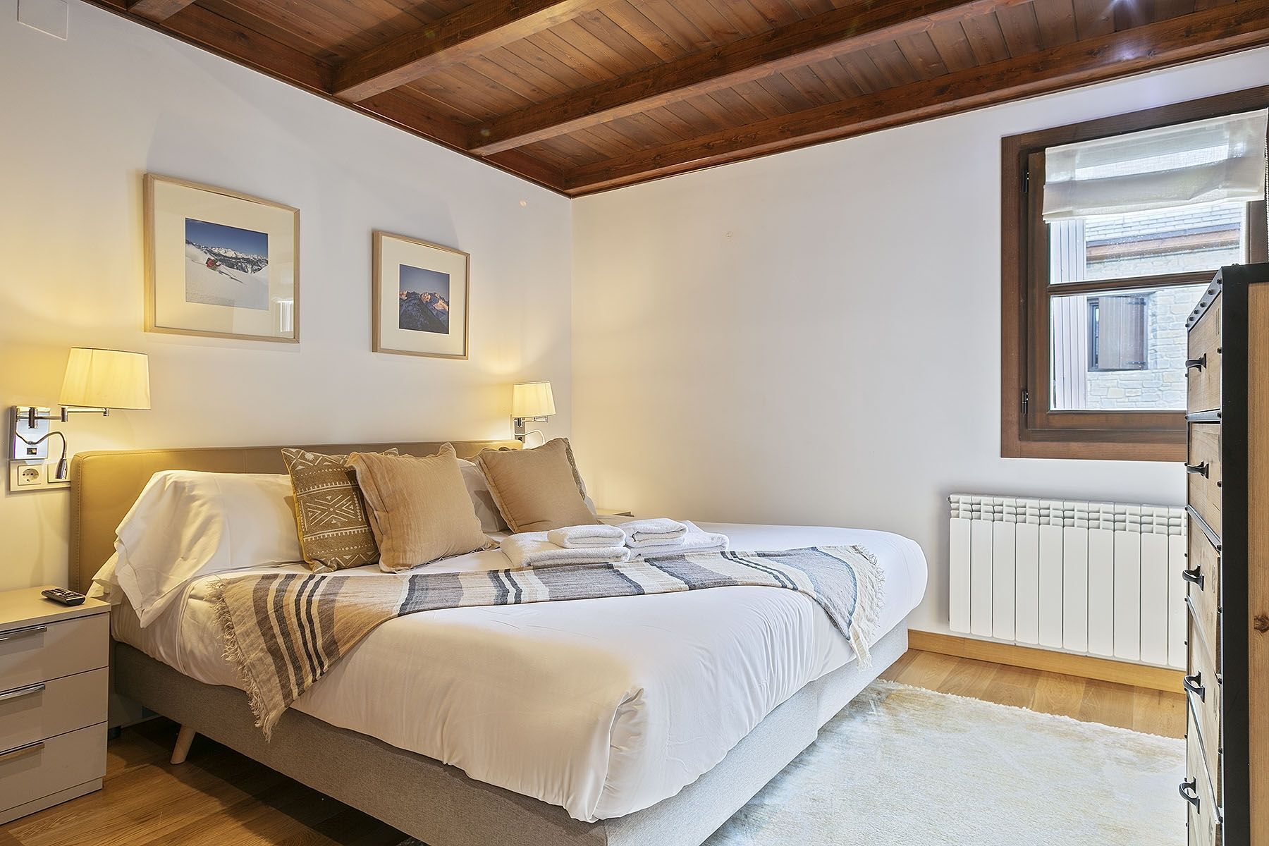 A bedroom with a wooden ceiling, a bed with white bedding, two framed pictures on the wall, and a window with blinds.