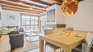 A spacious living room with a wooden ceiling, a dining area with a wooden table and chairs, and a large window providing natural light. - Luderna - Apartamento Val de Ruda B14 Paulet (Baqueira)