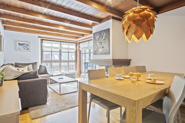 A spacious living room with a wooden ceiling, a dining area with a wooden table and chairs, and a large window providing natural light. - Luderna - Apartamento Val de Ruda B14 Paulet (Baqueira)