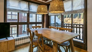 Et spiseområde med et træbord og stole, store vinduer med gardiner og et fjernsyn på et skab.