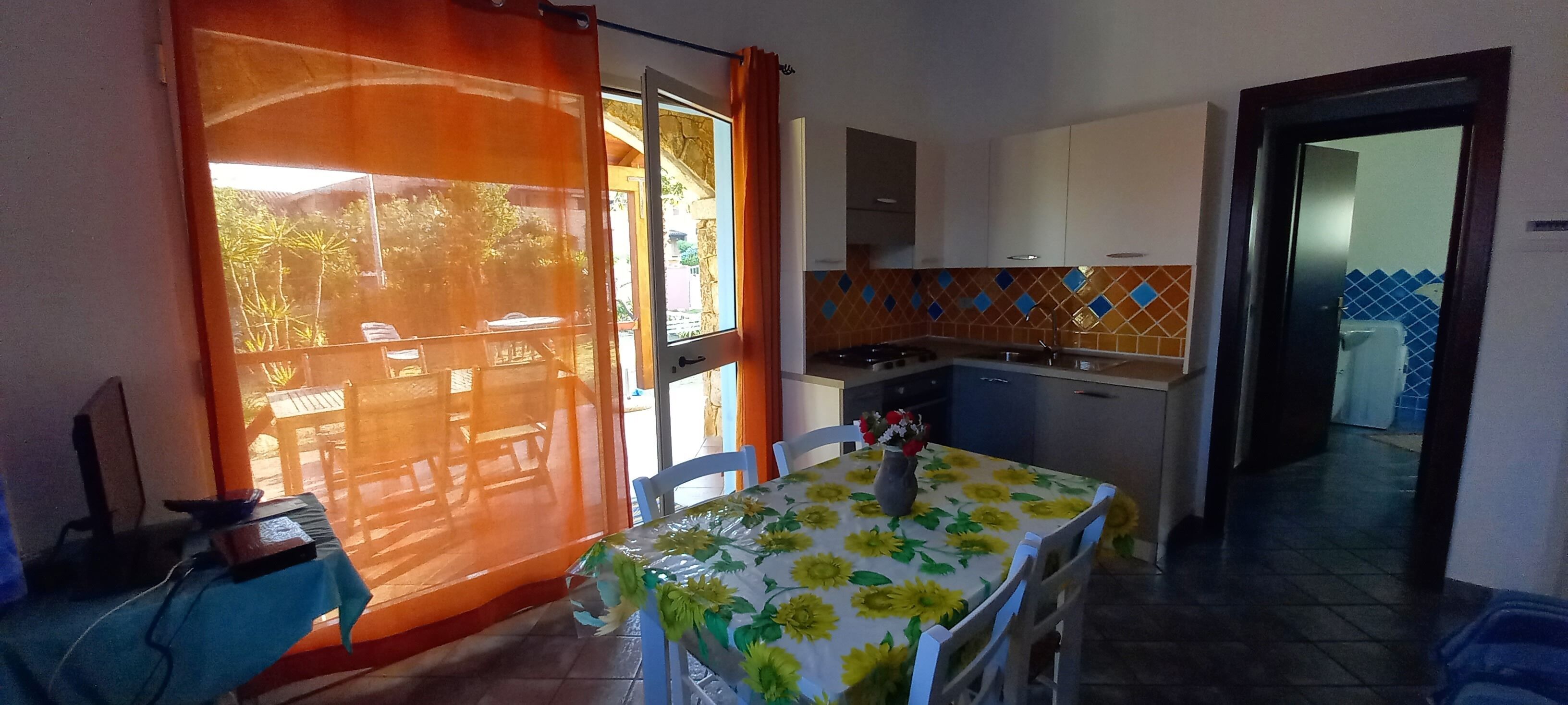 Una cocina compacta con zona de comedor, una mesa con mantel de girasol y vista al exterior a través de una ventana con cortina.