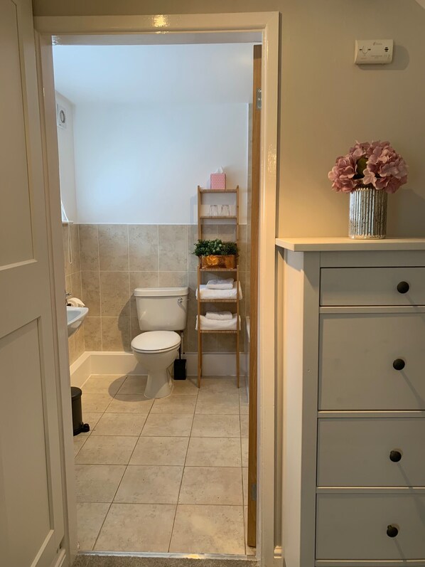 A bathroom with a toilet, sink, and shower. There is a shelf with towels and a vase with flowers. - Bakers Cottage (Telford)