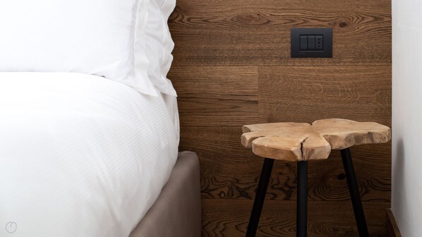 Una cama bien hecha con sábanas blancas, una mesita de noche de madera con un interruptor de luz moderno y un taburete sencillo de madera.