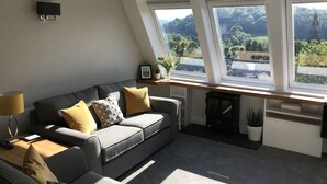 Una sala de estar moderna con un sofá gris, cojines amarillos y vistas al verdor a través de amplios ventanales.