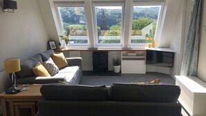 Una sala de estar moderna con un sofá, una mesita y un televisor. Tres ventanas ofrecen vistas al verdor exterior.