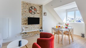 Un salon moderne avec un mur en pierre, un téléviseur à écran plat, un coin repas avec une table et des chaises, et une grande fenêtre offrant une vue sur la ville.