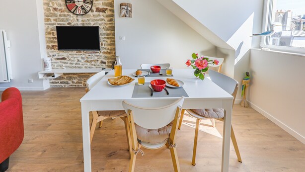 Un coin repas avec une table blanche dressée pour le petit-déjeuner, un mur en pierre avec un téléviseur et une fenêtre donnant sur des immeubles.