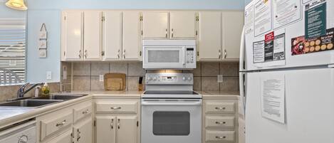 A kitchen with white cabinets, a microwave, an oven, and a refrigerator. There are wall-mounted papers and a hanging light fixture.