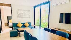 A modern living room with a sofa, coffee table, and dining table with chairs. There is a TV mounted on the wall and a window with a view of greenery. - 1 Bedroom apartment with views to green area and hamacs, Gym, Rooftop Pool (Playa del Carmen)