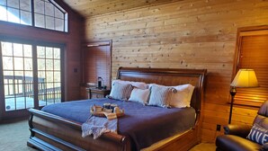 Ein großes Bett mit Holzkopfende, ein Nachttisch mit Lampe und Blick ins Freie durch große Fenster.