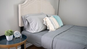 A neatly made bed with grey bedding, a white clock, and a small potted plant on a round table beside it. - Cute Home, Newly Remodeled, Great Location! (Grand Rapids)