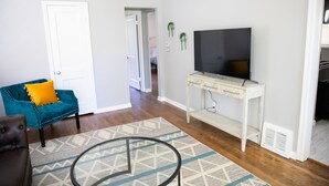A living room with a teal armchair, a brown sofa, a TV on a white console, and a patterned rug. - Cute Home, Newly Remodeled, Great Location! (Grand Rapids)