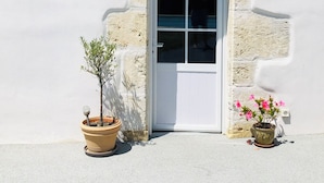 Un bâtiment à la façade blanche, doté d’un toit en tuiles et d’une porte blanche. On y trouve deux plantes en pot, l’une avec un petit arbre et l’autre avec des fleurs roses.