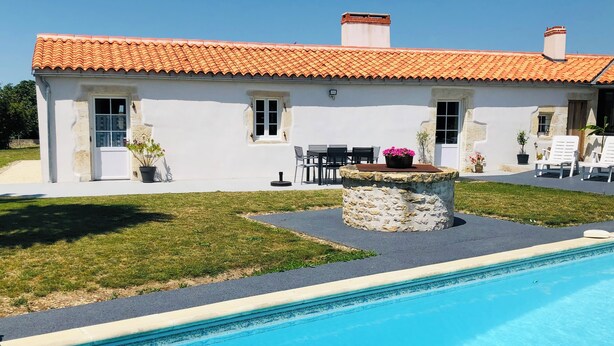 Un bâtiment blanc avec un toit en tuiles, un pilier en pierre surmonté d’un pot de fleurs et une piscine.