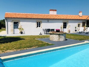 Un bâtiment blanc avec un toit en tuiles, un pilier en pierre surmonté d’un pot de fleurs et une piscine.