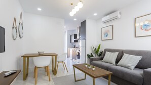Una sala de estar moderna con un sofá gris, una mesa de centro de madera y sillas blancas. Hay una mesa de comedor con un tazón y un control remoto sobre una mesita auxiliar. Una planta está en el suelo, y hay obras de arte en las paredes.