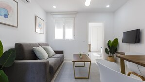 Un salón moderno con un sofá gris, una mesa de centro de madera, un sillón blanco y un televisor en la pared.