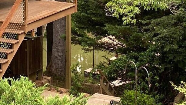 Une terrasse en bois avec un escalier menant à un deuxième niveau, entourée d’une végétation luxuriante et bordée par un plan d’eau.
