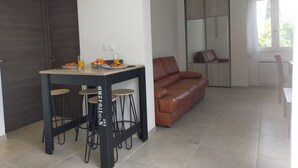 A modern living room with a brown leather sofa, a dining table with stools, and a large window with curtains. - T2 R+1 for 2/4 pers, private garden and parking! (Nîmes)