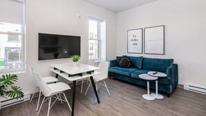 A modern living room with a white dining table, blue sofa, and a flat-screen TV mounted on the wall. - Amazing and Perfectly Located Flat by Denstays (Montréal)