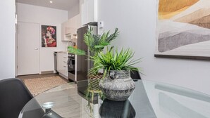 Een moderne keuken met witte kasten, een koelkast en een magnetron. Er is een eettafel met een glazen blad en een decoratieve plant in een pot met een patroon.