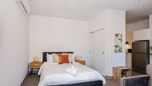 Een moderne hotelkamer met een groot bed, een houten nachtkastje, een klein tafeltje, een stoel en een kitchenette op de achtergrond.