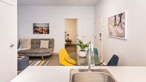 Una sala moderna con un sofá gris, un sillón amarillo, una zona de cocina con un fregadero y una mesa de comedor.