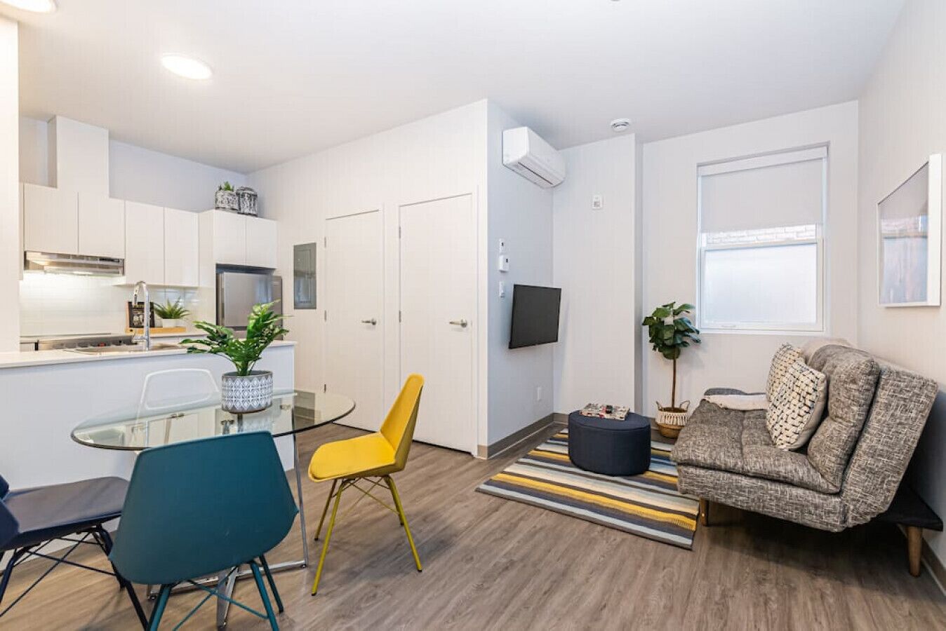 Un appartement moderne comprenant une cuisine, un coin repas et un salon. Il est équipé d’une table à manger en verre, d’une chaise jaune, d’un canapé gris et d’un téléviseur fixé au mur.
