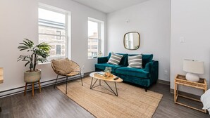 A modern living room with a teal sofa, a rattan chair, a coffee table, and a potted plant. - Brand New and Perfectly Located Flat in Le Plateau by Denstays (Montréal)