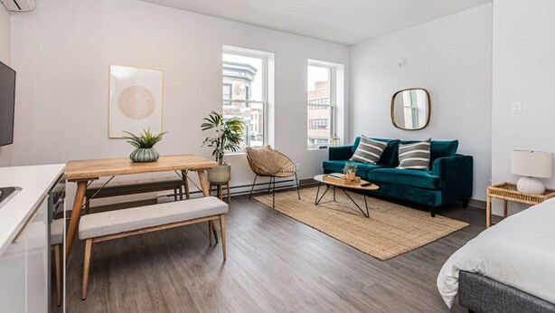 A modern living room with a wooden dining table, a teal sofa, and a large window providing a view of the city. - Brand New and Perfectly Located Flat in Le Plateau by Denstays (Montréal)