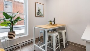 Una habitación pequeña y moderna con ventana, un escritorio con dos taburetes, una planta en maceta y un cuadro en la pared.