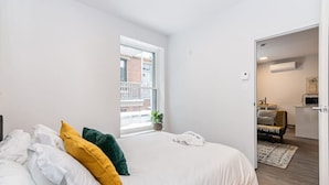 Un dormitorio con una cama grande, una ventana que ofrece vistas a un edificio, una planta pequeña y una puerta que da a un baño.