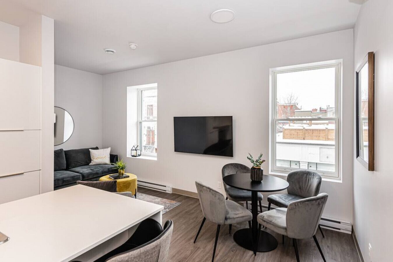 A modern living room with a sofa, a dining table, and chairs. There is a flat-screen TV mounted on the wall, and large windows providing natural light.